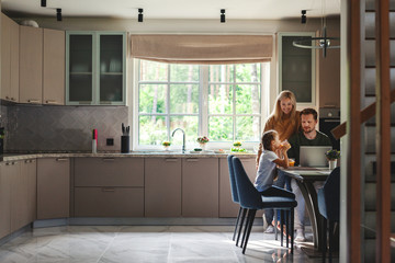 Middle aged man working on laptop computer in domestic kitchen surrounded by cute little daughter drinking juice and beautiful loving wife - Powered by Adobe