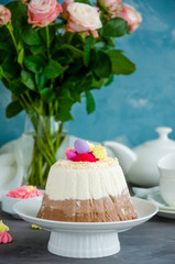 Easter cake. Cottage cheese Easter Paskha Three chocolates with coconut and candy eggs on top on a white plate on a concrete background with flowers. Vertical orientation. Copy space.