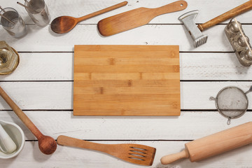 Drewniana deska kuchenna leży na białym stole. Na około leżą stare stylowe drewniane naczynia kuchenne tworzące tło. - obrazy, fototapety, plakaty