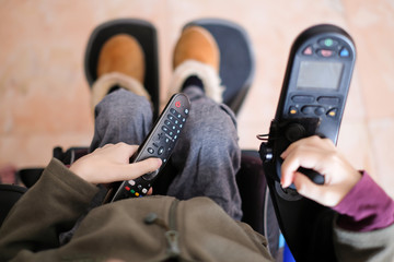A disabled person sitting in her electric wheelchair chilling while holding the tv control watching television at home during social distancing