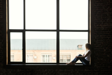 a young girl sits at home on the windowsill and looks out the window