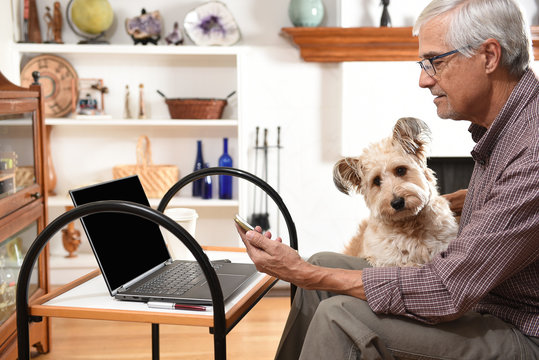 Mature man working from home with his laptop and cell phone and his pet dog on his lap, during the COVID-19 stay at home restrictions.