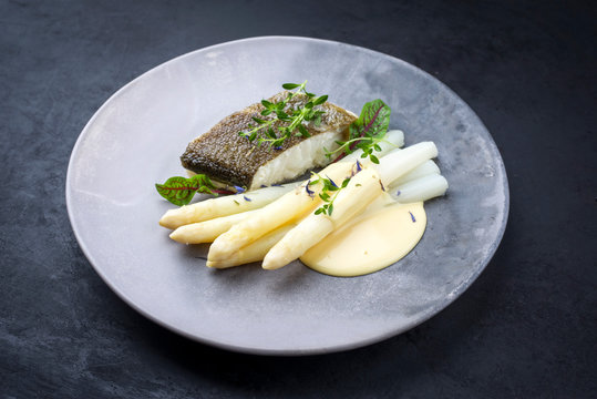 Gebratenes Gourmet Skrei Kabeljau Fisch Fillet mit wei&szlig;en Spargel und Sauce Hollandaise angeboten als closeup auf einem Modern Design Teller mit Textfreiraum