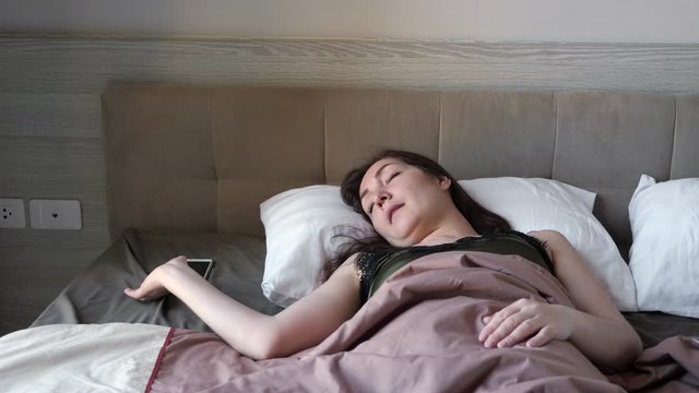 awaking woman looks at smartphone ringing wake-up call in large bed with beige blanket in hotel room in early morning