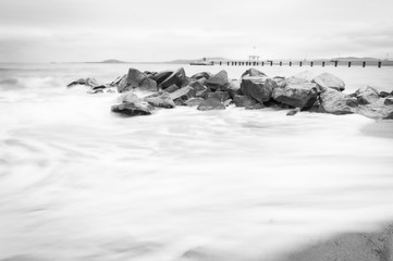 Rocks on the beach with silky sea water and Burgas bridge in the background. Black and white Fine art image of coast in Bulgaria. Travel destination 2020.