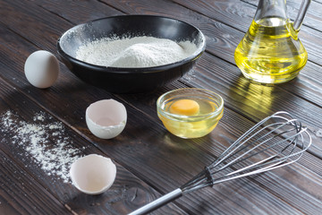 Sieve on black bowl with flour. Glass bottle with oil, eggs, eggshell, broken egg in plate, metal whisk, wooden spoon with flour.