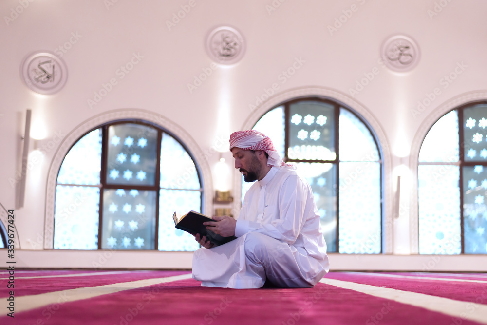 Wall mural Young Arabic Muslim man reading Koran and praying. Religious muslim man reading holy koran inside the mosque.