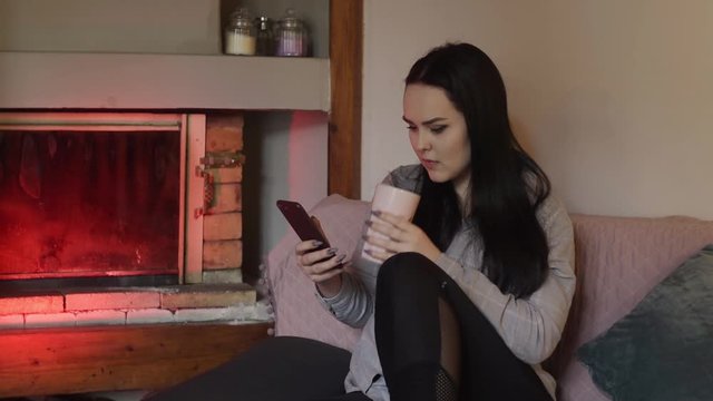 The brunette is sitting at home and looking at the phone, the girl is chatting, playing games, smiling. The girl sits dma near the fireplace, all the time on the phone. 