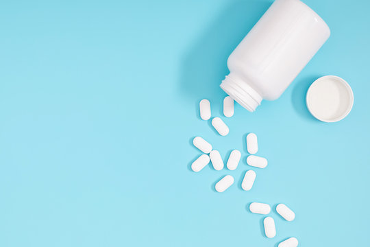 White pills spilled out of white bottle on light blue background. Minimal medical concept. Flat lay, top view, copy space