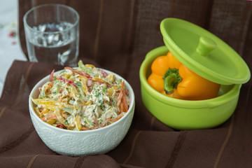 Decorated with lettuce, side view, selective focus . Orange pepper in a green bowl . Water in a glass. Brown tablecloth. Salad with ham, bell pepper, mushrooms, pickles, white sauce, on a white plate.
