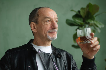 Side view of senior man holding glass of whisky or brandy and thinking about something in loft style room with light green walls and houseplants on background. Alcohol addiction