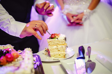 Obraz na płótnie Canvas Bride and a groom is cutting their rustic wedding cake on wedding banquet. The bridegroom eats a cake. The bridegroom takes a piece of wedding cake covered with icing and decorated with pink roses
