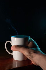 Taza de café blanca y caliente saliendo humo en la oficina de casa y mano que la sujeta para tomar  un sorbo 