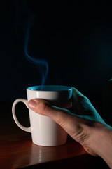 Taza de café blanca y caliente saliendo humo en la oficina de casa y mano que la sujeta para tomar...