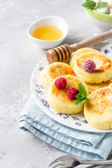 Cottage cheese pancakes, syrniki, curd fritters with honey, raspberry and mint. Gourmet breakfast. Light grey concrete background. Selective focus. 
