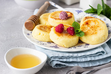 Cottage cheese pancakes, syrniki with honey, raspberry and mint on ceramic plate and homemade cottage cheese in a bowl. Healthy breakfast or lunch.