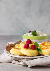 Cottage cheese pancakes, syrniki with honey, raspberry and mint on ceramic plate and homemade cottage cheese in a bowl. Healthy breakfast or lunch.
