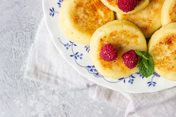 Cottage cheese pancakes, syrniki, curd fritters with honey, raspberry and mint. Gourmet breakfast. Light grey concrete background. Selective focus. 