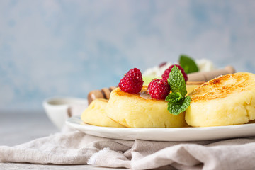 Cottage cheese pancakes, syrniki with honey, raspberry and mint on ceramic plate and homemade cottage cheese in a bowl. Healthy breakfast or lunch.