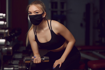 The girl in a protective mask is engaged in dumbbells. Protective masks against virus infection. Training during quarantine in the gym. Coronavirus (COVID-19) protection concept.