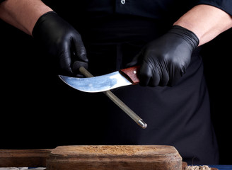 chef in a black shirt and black latex gloves sharpen a kitchen knife on an iron sharpener with a handle