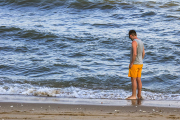 Fototapeta na wymiar young man wearing yellow swimming shorts and a gray t-shirt stands on the shore and looks at the ocean, left side view
