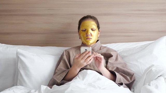 brunette girl with golden face mask uses modern smartphone to surf social nets lying in soft bed in comfortable hotel room