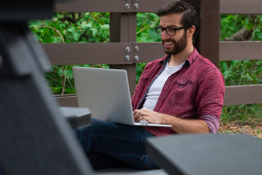 A Young Bearded Hipster Man With Glasses Sits Against A Pavilion Pillar Outside On A Cloudy Covered Day Near A Walking Path Working Remotely On His Laptop Trying To Socially Distance Himself.