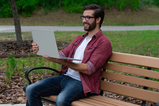 A Young Bearded Hipster Man With Glasses Sits On A Park Bench Outside On A Cloudy Covered Day Near A Walking Path Working Remotely On His Laptop Trying To Socially Distance Himself