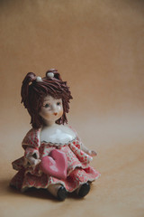 Small statue of little lady with pink heart and brown hair. Greeting card with flower girl. Close up picture of small toy. Brown background. Accessories for home. Vintage doll. 