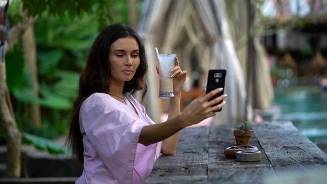 Young beautiful woman with drink taking selfie photo with cellphone in park cafe