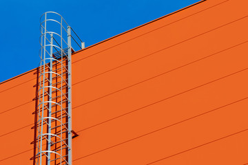 A fragment of a orange modern facade of an industrial building, warehouse or shopping center with a long metal staircase against the blue sky. Staircase to heaven symbol