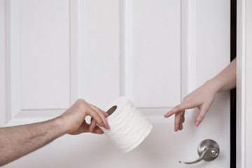 A mans hand passing a roll of toilet paper to a womans hand coming out of a door.