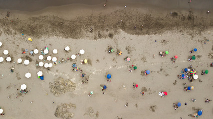 Aerial view on the beach with colourful umbrellas and tourists enjoying their time during weekend at the Peruvian coast. Rocky coast, brown sand, green waters, waves.