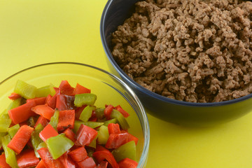 Cooked casserole ingredients of hamburger meat and chopped red and green bell peppers and in bowls on yellow background