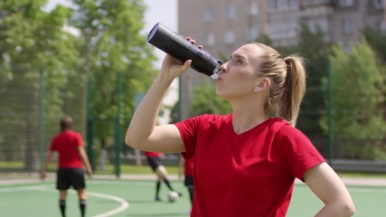 Young female soccer player in uniform standing on field and drinking water from sports bottle while taking a break during training