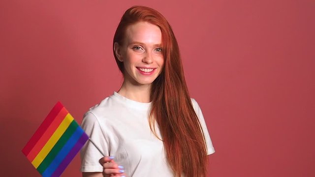 redhaired ginger woman in pink studio background waving rainbow colorfull flag