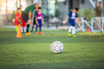 Fototapeta premium Old soccer ball on green artificial turf with blurry soccer team training. Blurry kid soccer player jogging between marker cones and control ball with soccer equipment in football academy.