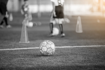 Black and white background image. Old soccer ball on green artificial turf with blurry soccer team...