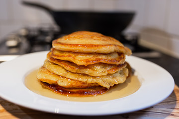 Fresh homemade classic pancakes with maple syrup in a white plate on a wooden board with a pan in the background.