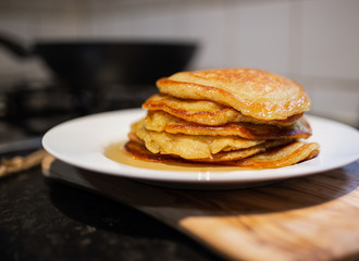 Fresh homemade classic pancakes with maple syrup in a white plate on a wooden board with a pan in the background.