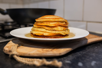 Fresh homemade classic pancakes with maple syrup in a white plate on a wooden board with a pan in the background.