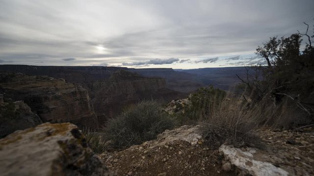 A beautiful wide sunset motion timelapse of the Grand Canyon. The camera tracks past foreground rocks and vegetation before finishing the move as the sun sets below the horizon and the sky lights up w
