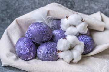 Spotted painted purple eggs of a chicken. Colorful easter eggs. Colored eggs purple on an eco canvas bag with a feather and cotton bolls. Purple eggs on a gray background. Happy easter