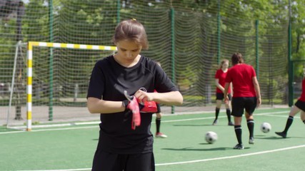 Young girl standing on outdoor soccer field and putting on goalkeeper gloves while her female teammates exercising in the background