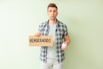 Young man holding paper with text HEMORRHOIDS and toilet paper in blood on color background