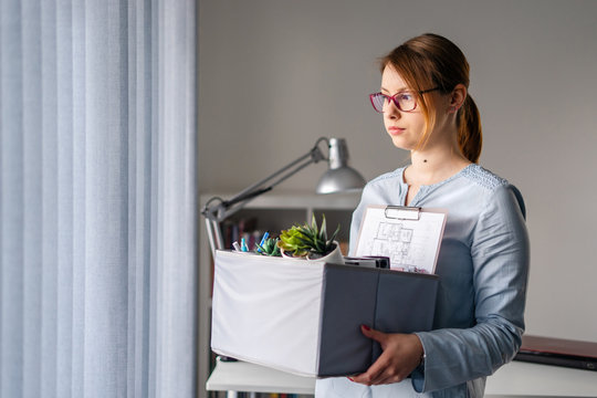 Young Adult Caucasian Woman Female Girl Standing By The Window At The Office Losing Her Job Holding Personal Items Things In Box Being Fired From Work Dismissed Due To Crisis Recession Quitting