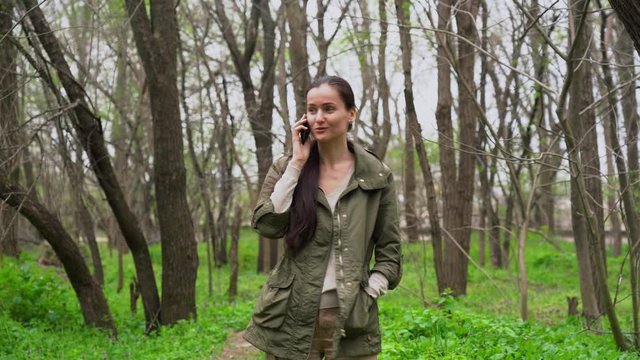 Young beautiful Caucasian girl with long brown hair in autumn warm jacket walks alone in the empty forest among trees with green grass, talk on the mobile phone, smile, laugh outdoor in cold morning.