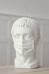 Plaster sculpture of Caesar's head stands on wooden table in medical mask. Protection against infections and coronavirus.