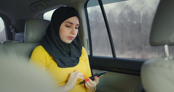 oung beautiful muslim woman in hijab in car passenger rear seat typing on cell phone with serious expression. Internet bank. Businesswoman working at smartphone in back seat of crowdsourced taxi